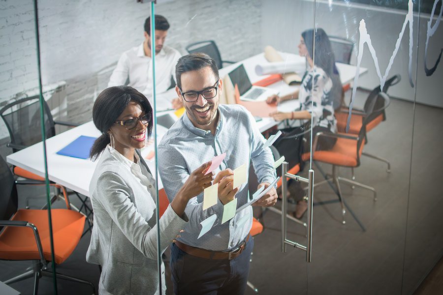 Employee Benefits - Two Employees Posting Sticky Notes on Glass Wall in Office