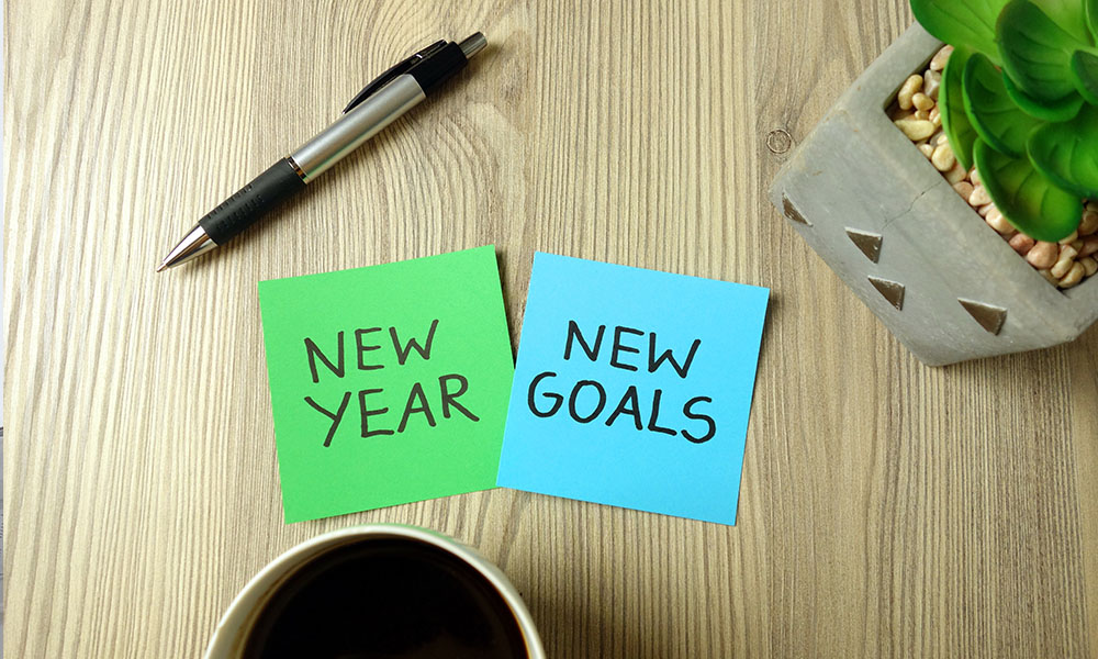 Blog - Coffee Cup, Pen, Plant, a Post-it Note with the Text New Year, and a Post-it Note with the Text New Goals on a Wooden Desk